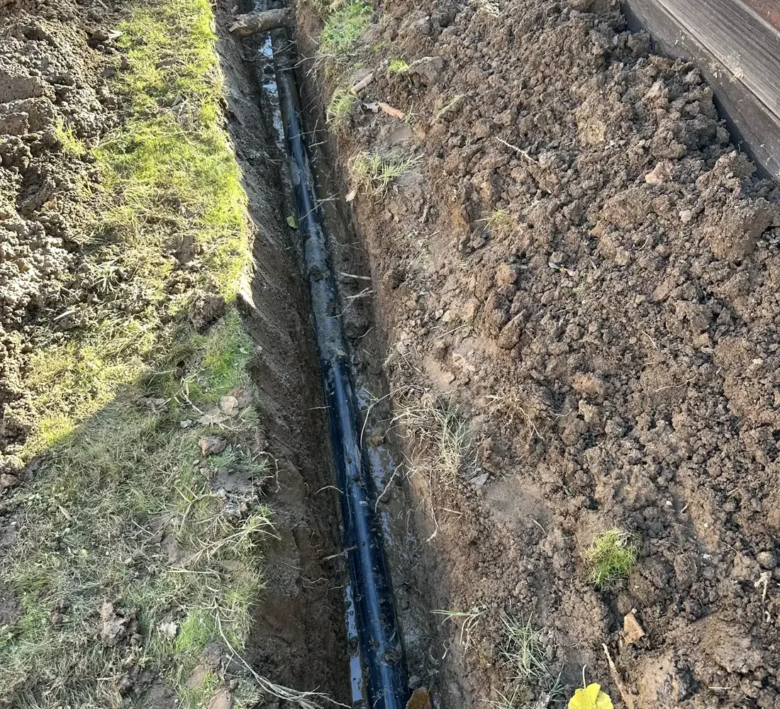 Long exterior trench with newly installed black sewer piping beside a residential structure.
